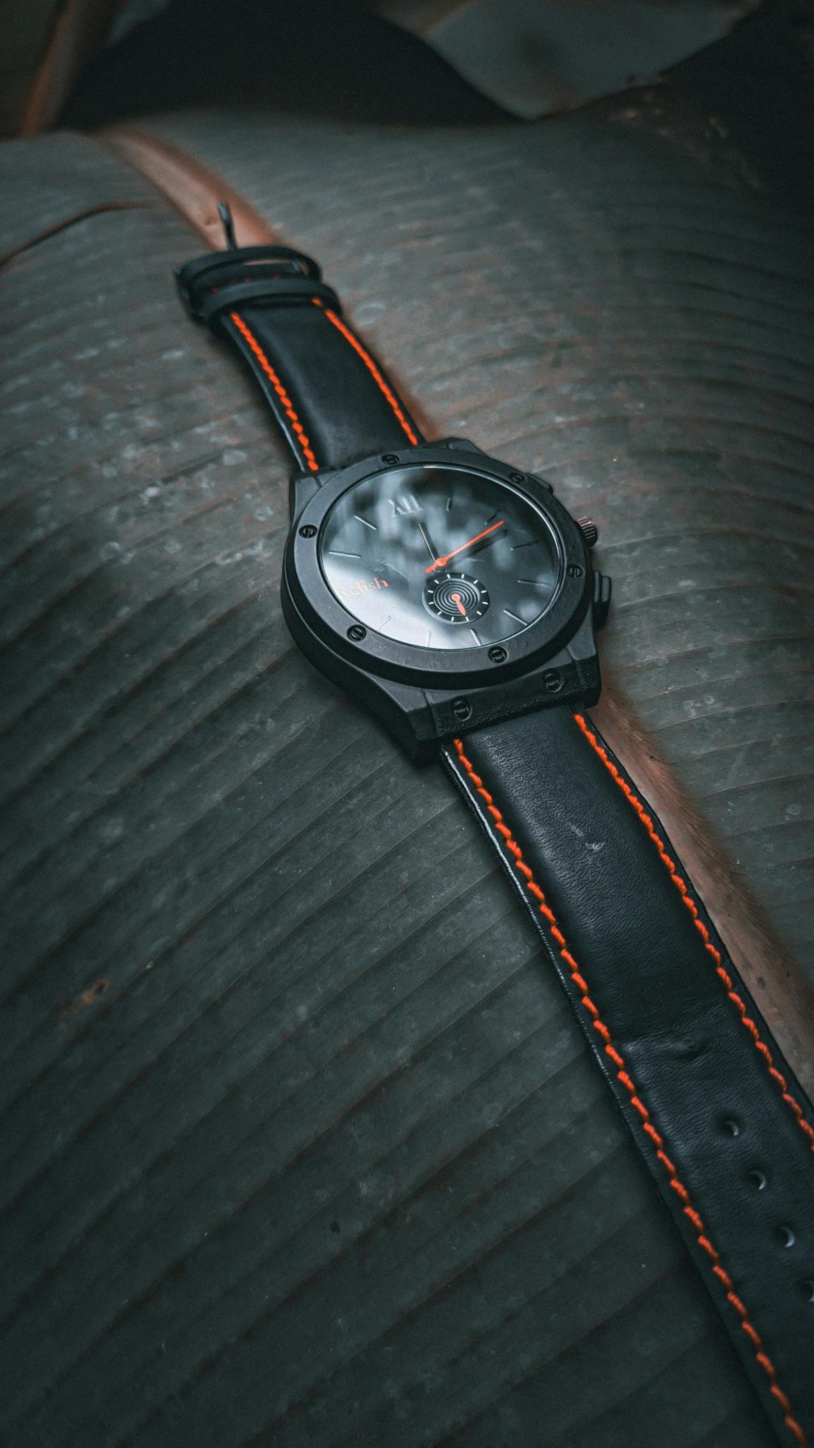 Sleek black wristwatch with orange accents lies on textured leather, conveying elegance.